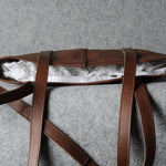 A brown leather shoulder bag with a long strap, shown open with white tissue paper inside, resting on a textured gray surface.