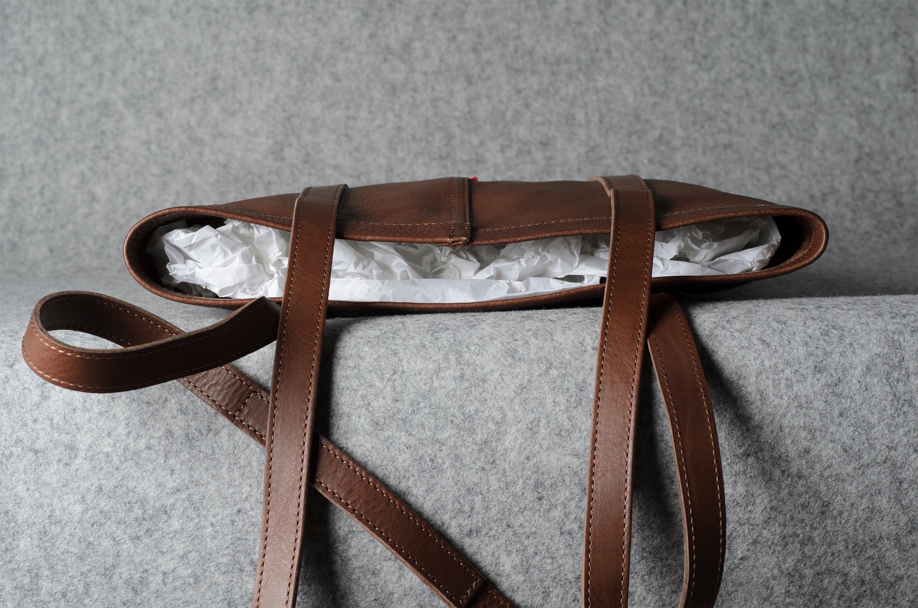 A brown leather shoulder bag with a long strap, shown open with white tissue paper inside, resting on a textured gray surface.