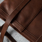 A close-up of a brown leather tote bag showing the pebbled texture, clean stitching, and a matching leather strap on a gray textured surface.