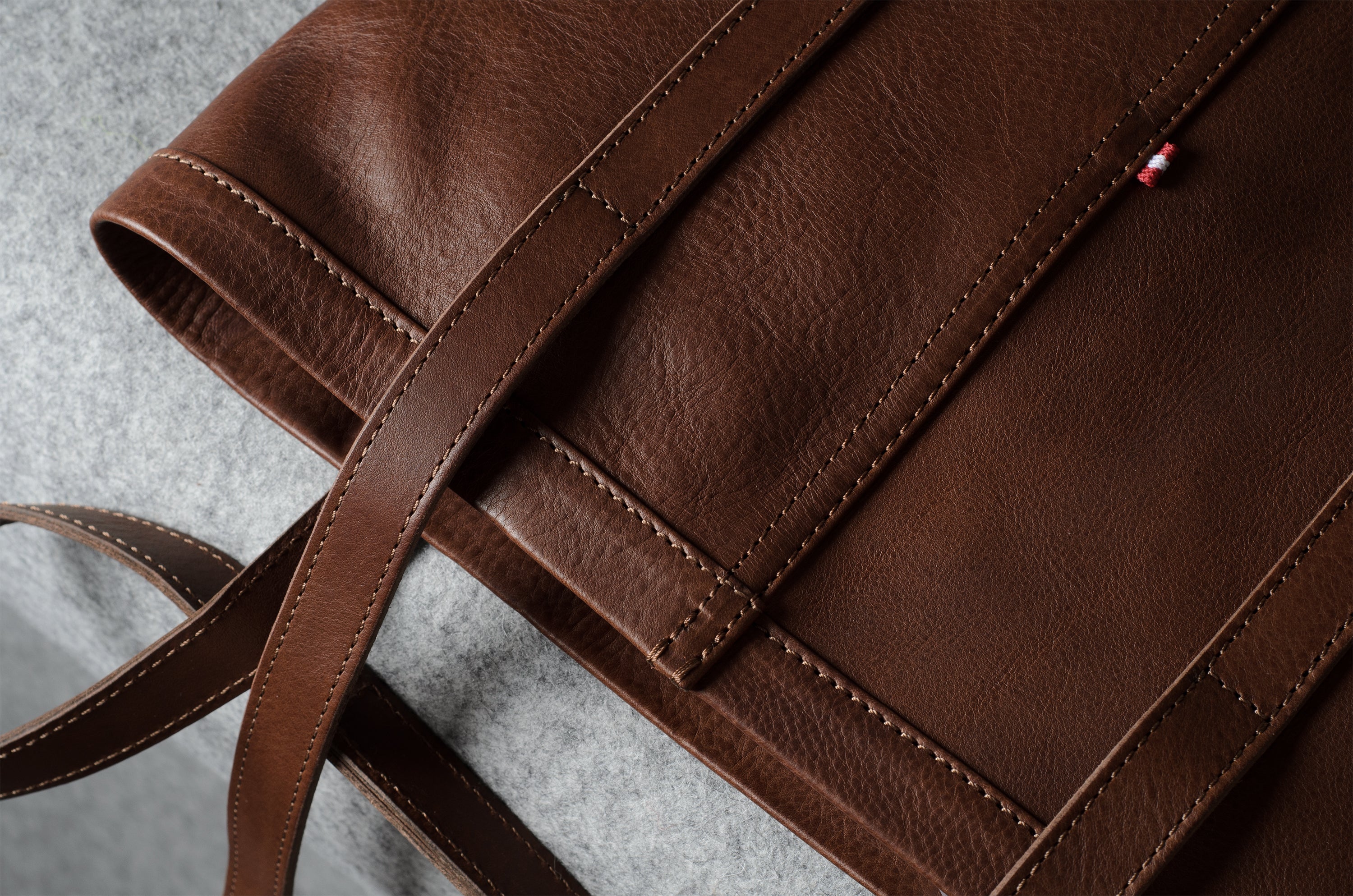 A close-up of a brown leather tote bag showing the pebbled texture, clean stitching, and a matching leather strap on a gray textured surface.