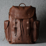 A brown leather backpack with a top flap, drawstring closure, and side pockets, photographed against a textured gray background.