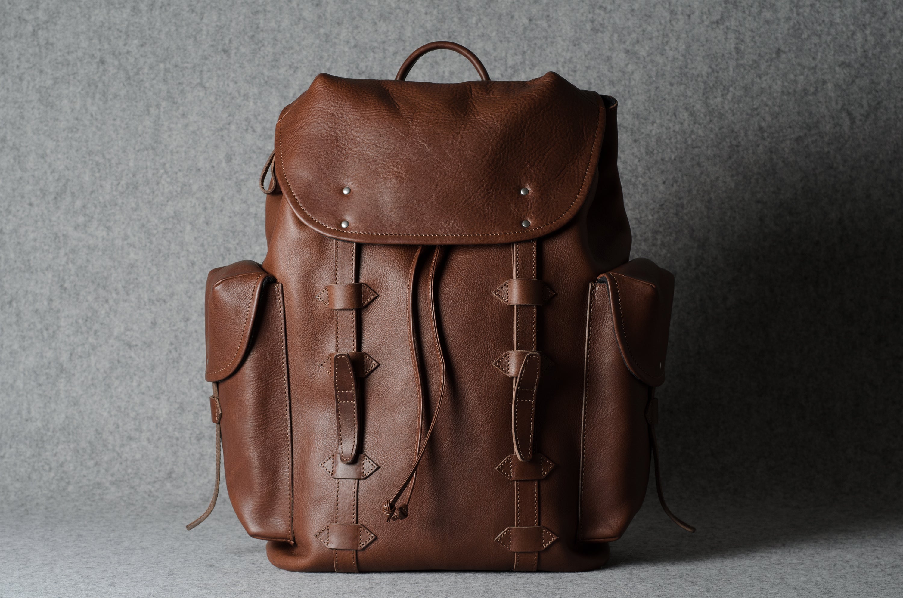 A brown leather backpack with a top flap, drawstring closure, and side pockets, photographed against a textured gray background.
