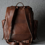 A brown leather backpack shown from the back, featuring dual top handles, side pockets, and adjustable shoulder straps, photographed against a textured gray background.