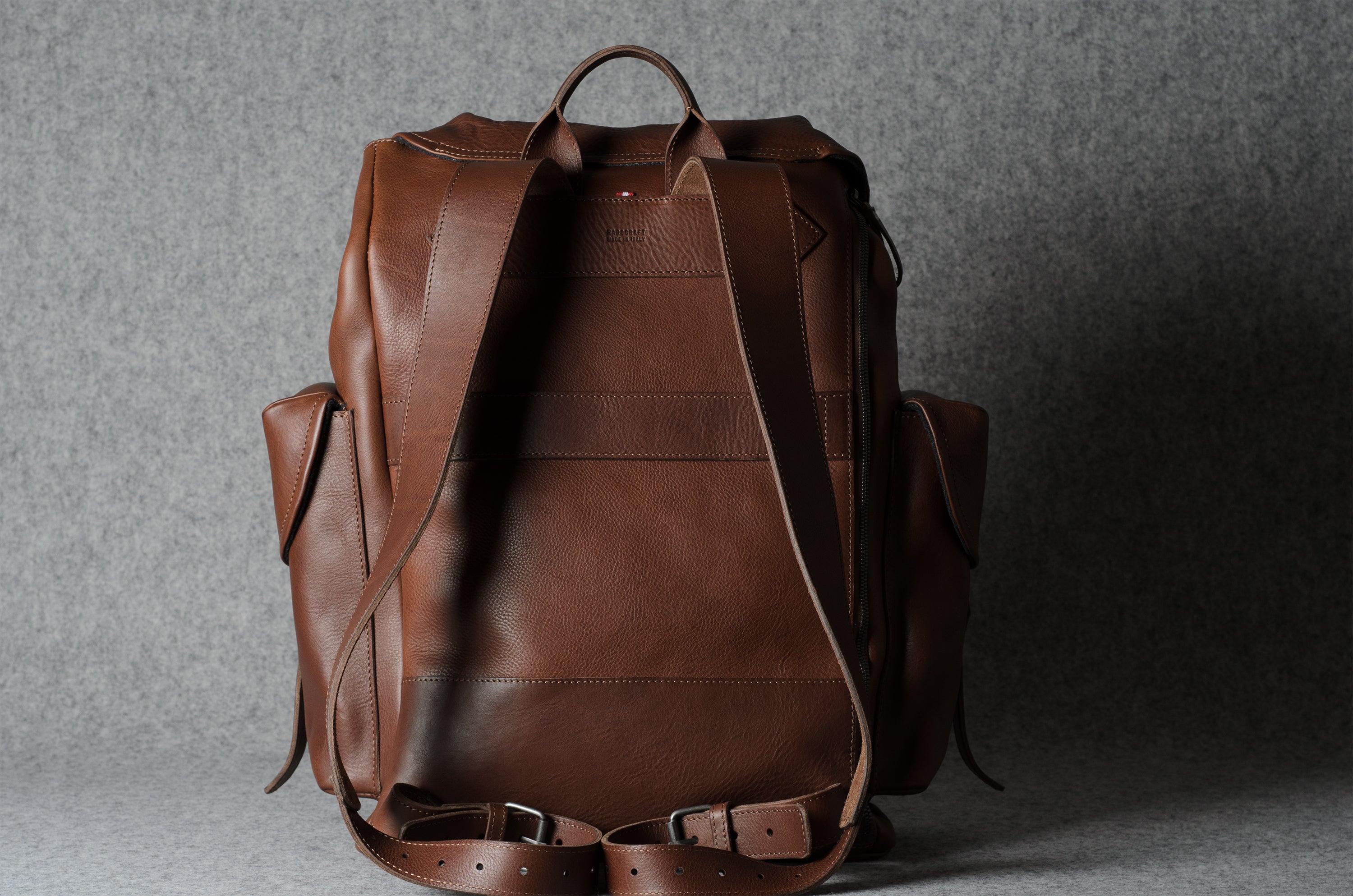 A brown leather backpack shown from the back, featuring dual top handles, side pockets, and adjustable shoulder straps, photographed against a textured gray background.
