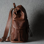 A brown leather backpack with a front flap pocket and adjustable shoulder strap, photographed against a textured gray background.
