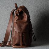A brown leather backpack with a front flap pocket and adjustable shoulder strap, photographed against a textured gray background.