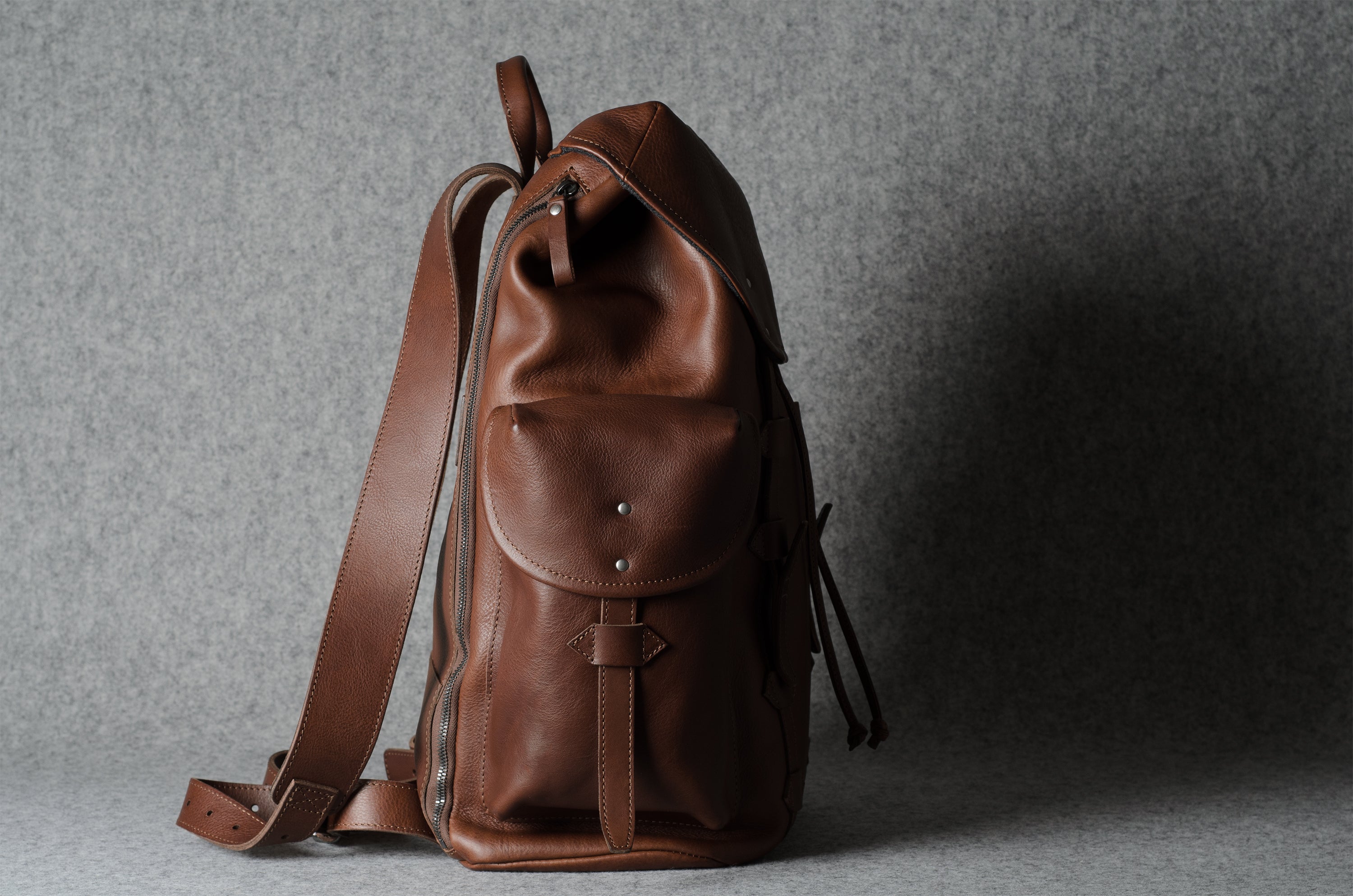 A brown leather backpack with a front flap pocket and adjustable shoulder strap, photographed against a textured gray background.