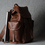 A brown leather backpack with a flap top, front pocket, and shoulder strap, shown standing upright against a gray textured background with a silver laptop tucked inside.