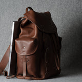 A brown leather backpack with a flap top, front pocket, and shoulder strap, shown standing upright against a gray textured background with a silver laptop tucked inside.