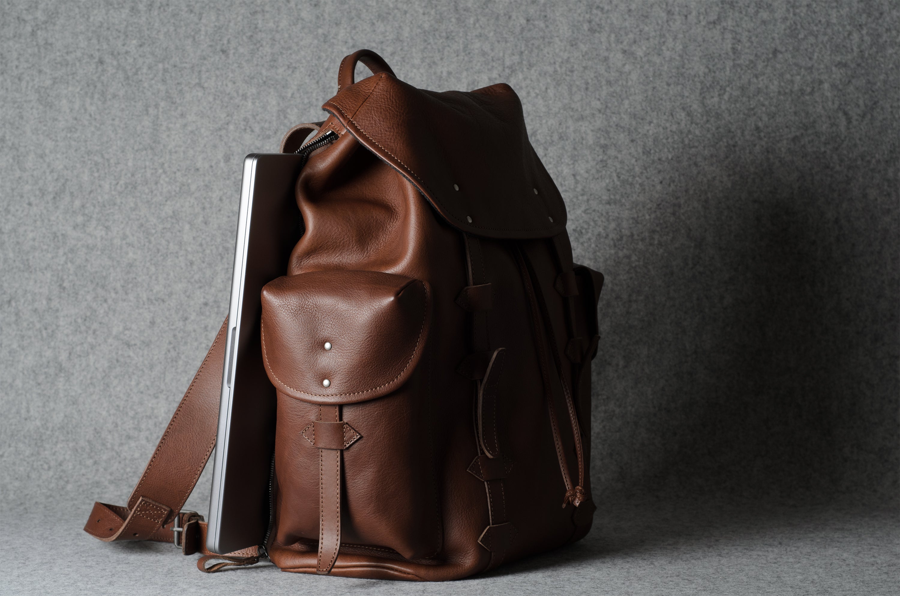 A brown leather backpack with a flap top, front pocket, and shoulder strap, shown standing upright against a gray textured background with a silver laptop tucked inside.
