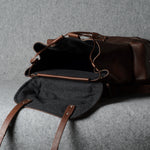 A dark brown leather duffel bag with an open flap showing a black interior, photographed on a textured gray fabric background.
