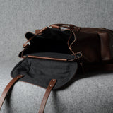 A dark brown leather duffel bag with an open flap showing a black interior, photographed on a textured gray fabric background.