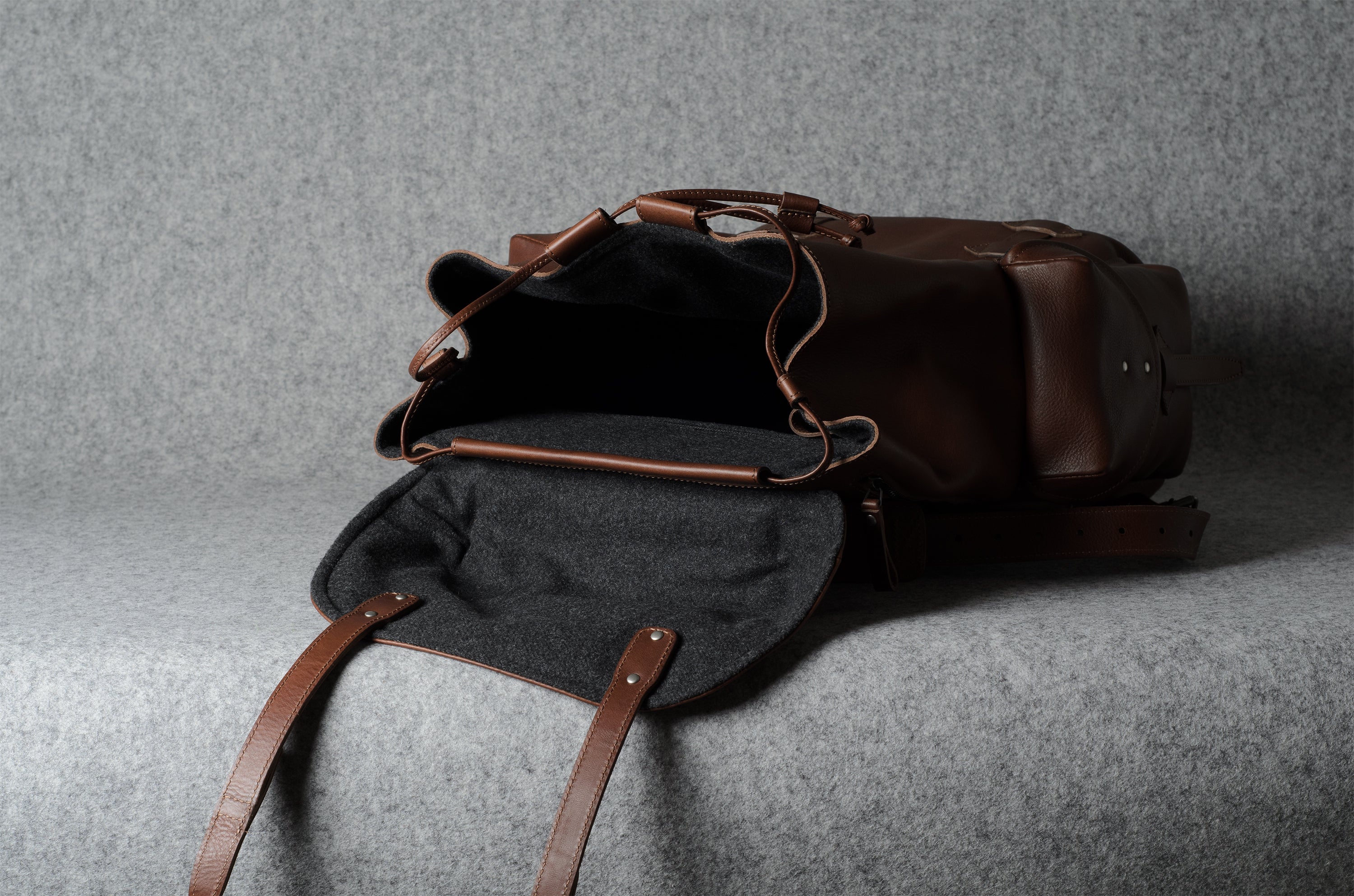 A dark brown leather duffel bag with an open flap showing a black interior, photographed on a textured gray fabric background.