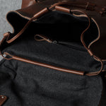 Close-up of a brown leather bag with an open flap showing a dark gray felt interior, zipper pocket, and leather trim on a gray textured surface.