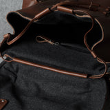 Close-up of a brown leather bag with an open flap showing a dark gray felt interior, zipper pocket, and leather trim on a gray textured surface.
