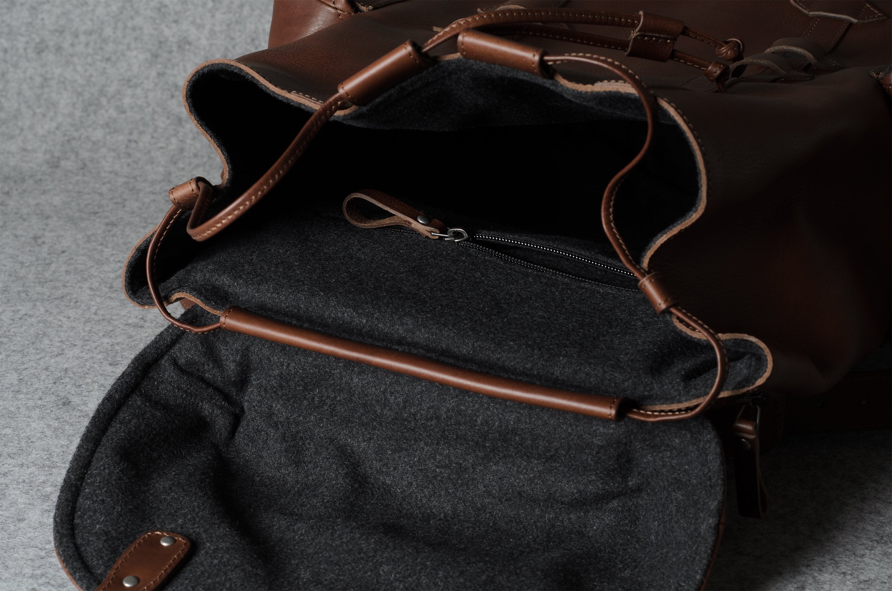 Close-up of a brown leather bag with an open flap showing a dark gray felt interior, zipper pocket, and leather trim on a gray textured surface.