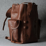 A brown leather backpack with a front pocket and buckle details, photographed against a textured gray background.