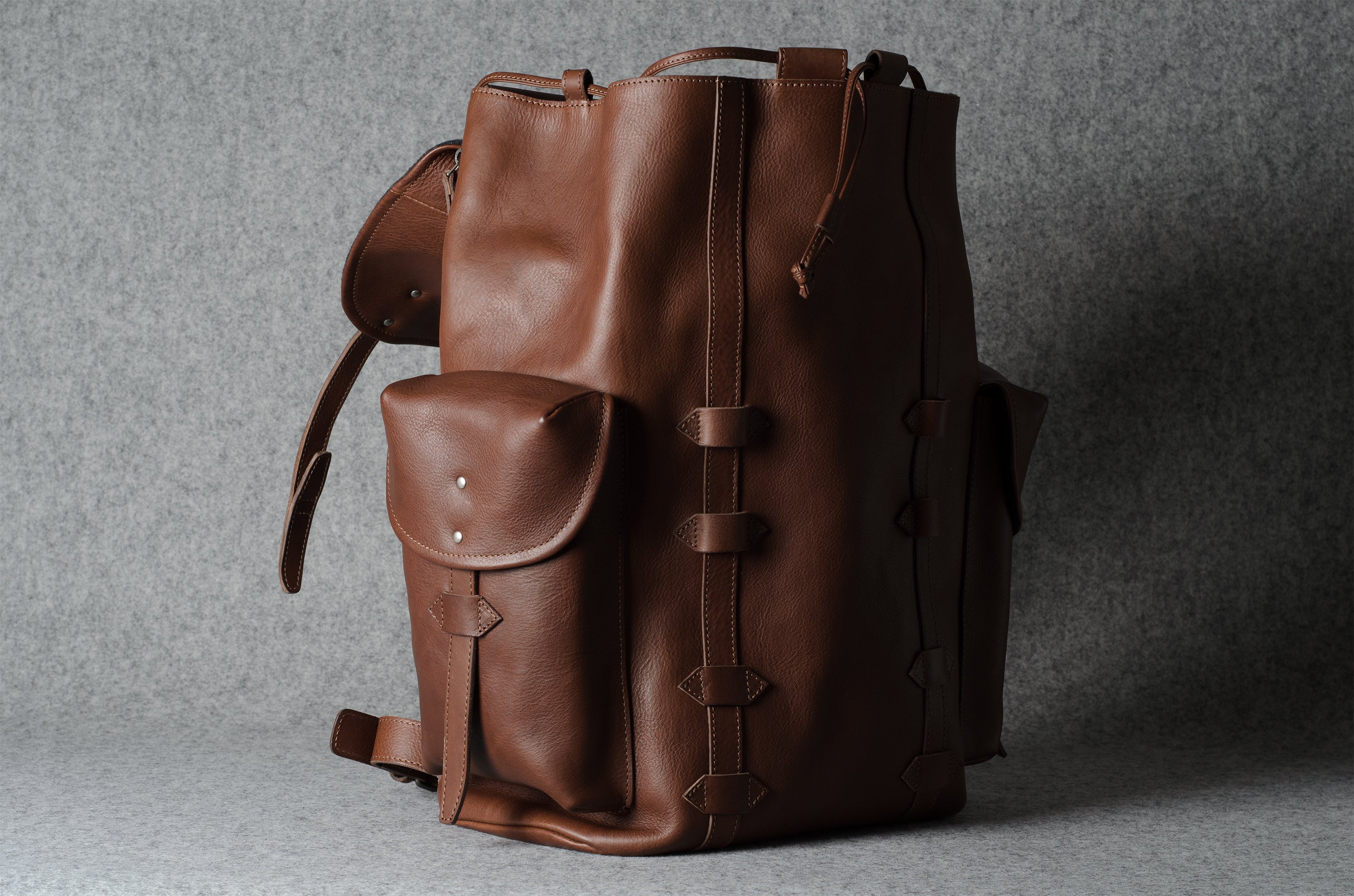 A brown leather backpack with a front pocket and buckle details, photographed against a textured gray background.