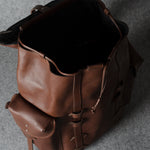 A brown leather backpack shown from an overhead angle, with its main compartment open and a smaller front pouch attached, set against a textured gray background.