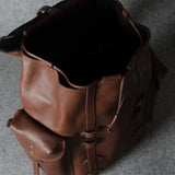 A brown leather backpack shown from an overhead angle, with its main compartment open and a smaller front pouch attached, set against a textured gray background.