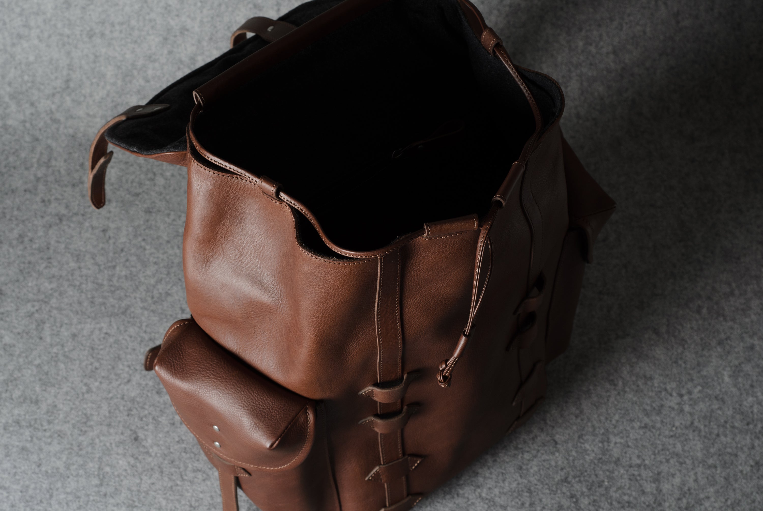 A brown leather backpack shown from an overhead angle, with its main compartment open and a smaller front pouch attached, set against a textured gray background.