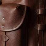 Close-up of a brown leather backpack showing a front flap pocket with silver rivets and a vertical strap closure on the side.