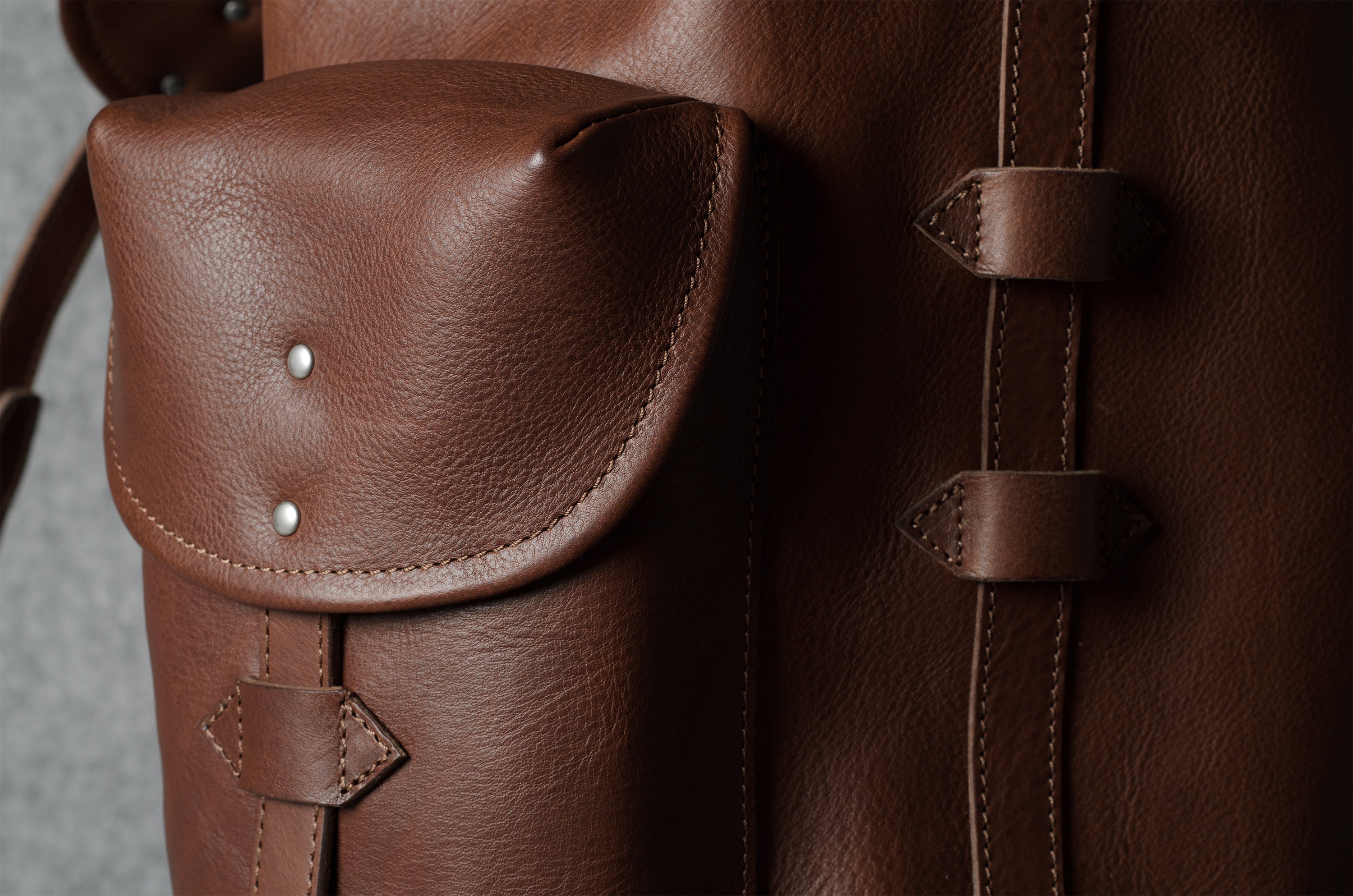 Close-up of a brown leather backpack showing a front flap pocket with silver rivets and a vertical strap closure on the side.