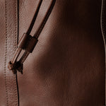 Close-up of a brown leather bag showing the textured leather surface, vertical stitching, and a matching leather drawstring with a small knot detail.