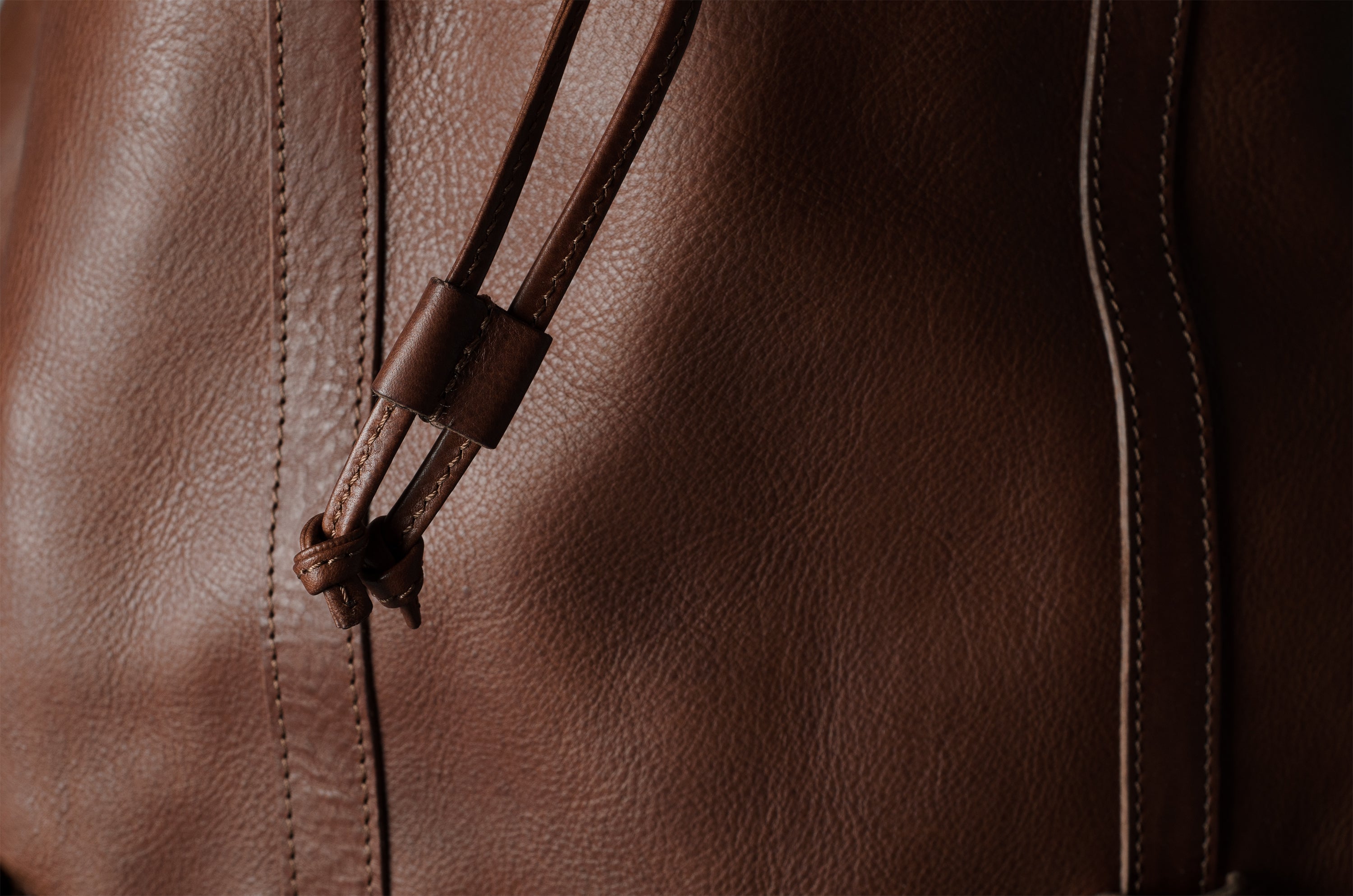 Close-up of a brown leather bag showing the textured leather surface, vertical stitching, and a matching leather drawstring with a small knot detail.