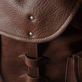 Close-up of a brown leather bag showing the flap, metal rivets, and strap details with a dark background.