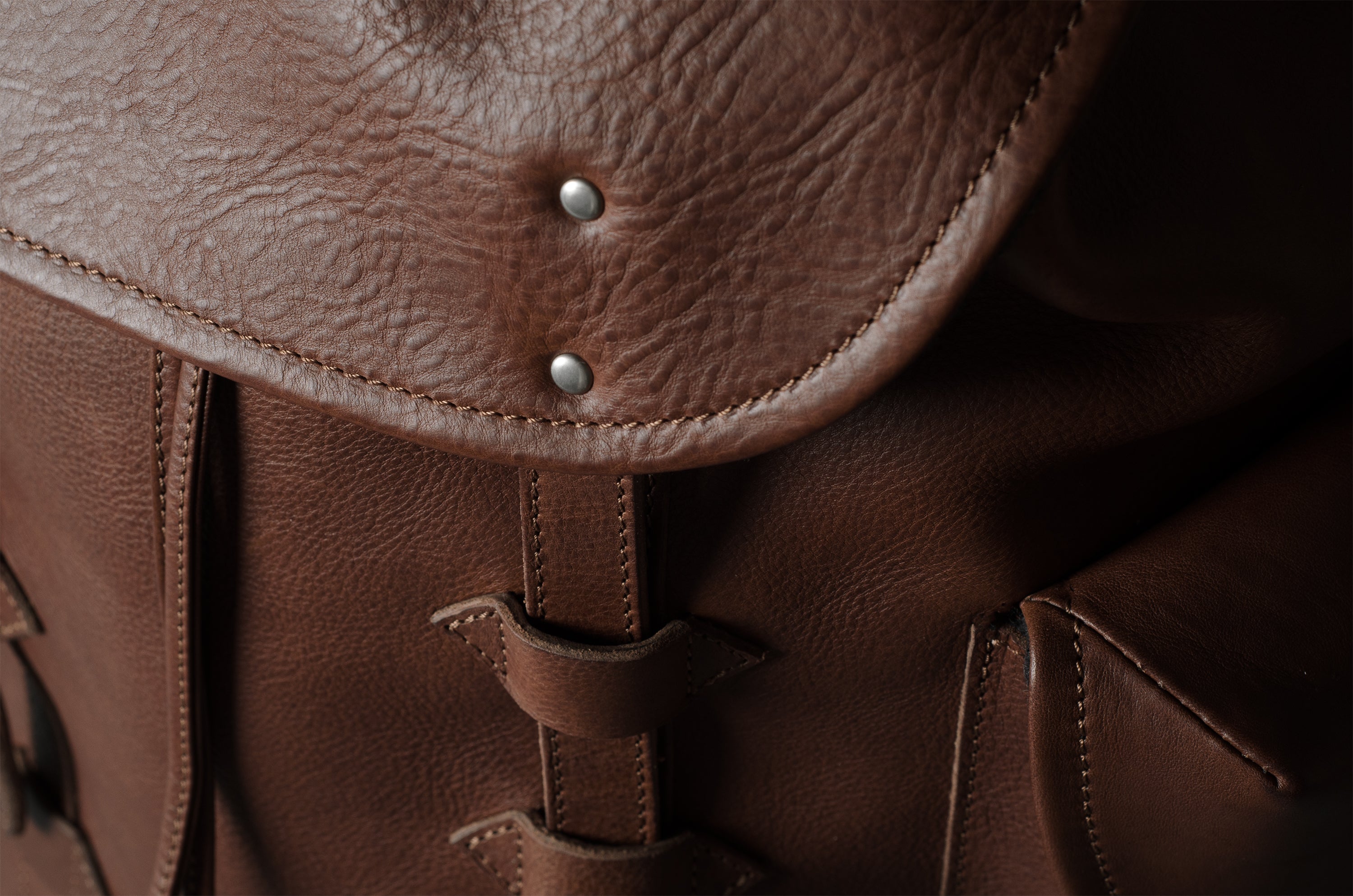 Close-up of a brown leather bag showing the flap, metal rivets, and strap details with a dark background.