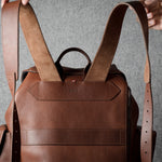 A brown leather backpack with dual shoulder straps and a top handle, held by hands against a textured gray background.