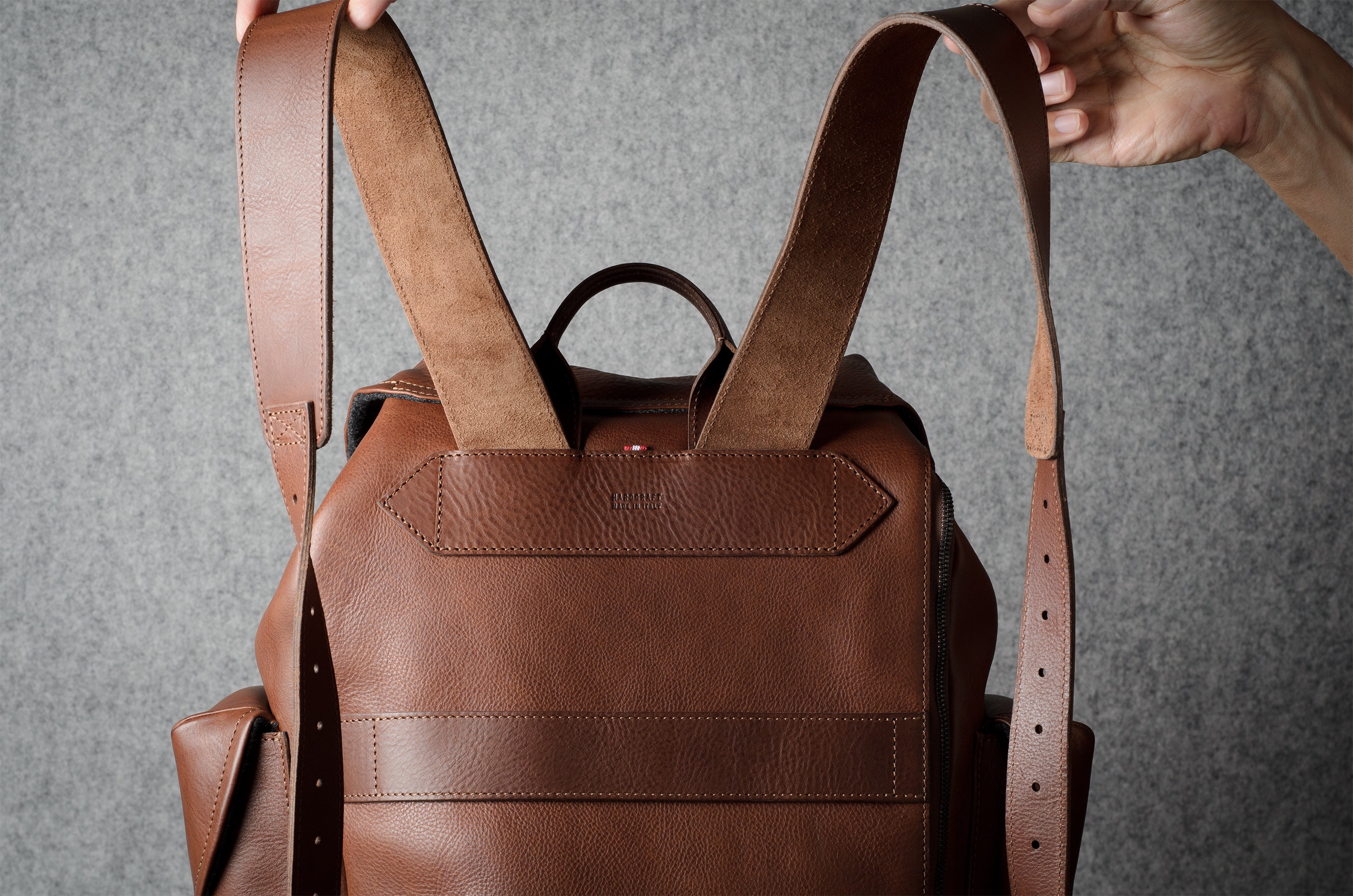 A brown leather backpack with dual shoulder straps and a top handle, held by hands against a textured gray background.