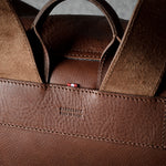 Close-up of a brown leather handbag showing the back panel with a stitched leather tab embossed with ‘HARDGRAFT MADE IN ITALY' and a small red brand tag, with suede-like handles rising above.
