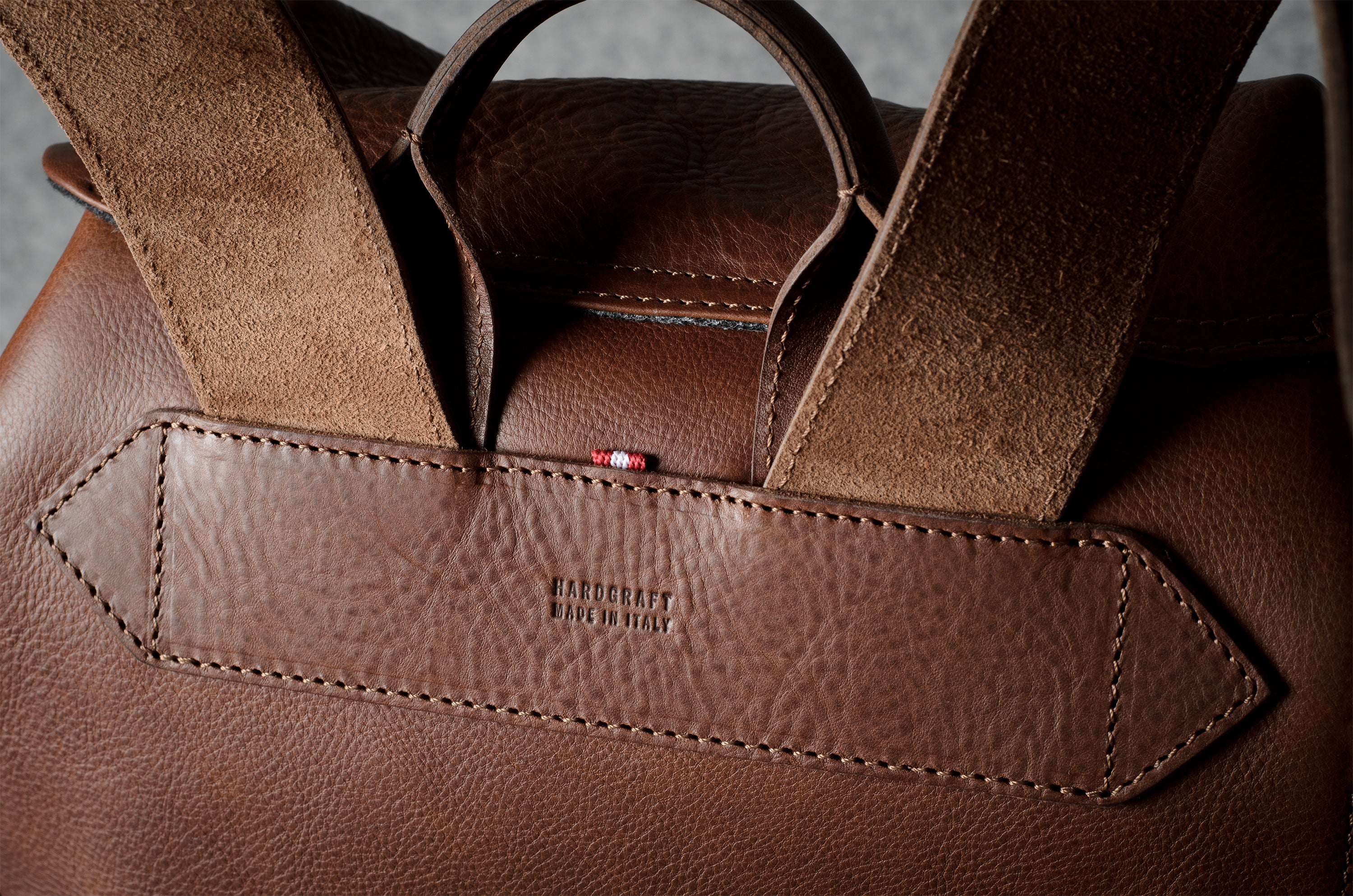 Close-up of a brown leather handbag showing the back panel with a stitched leather tab embossed with ‘HARDGRAFT MADE IN ITALY' and a small red brand tag, with suede-like handles rising above.