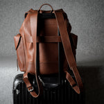 A brown leather backpack with wide adjustable shoulder straps, shown from the back resting on top of a black hard-shell suitcase against a textured gray studio background.