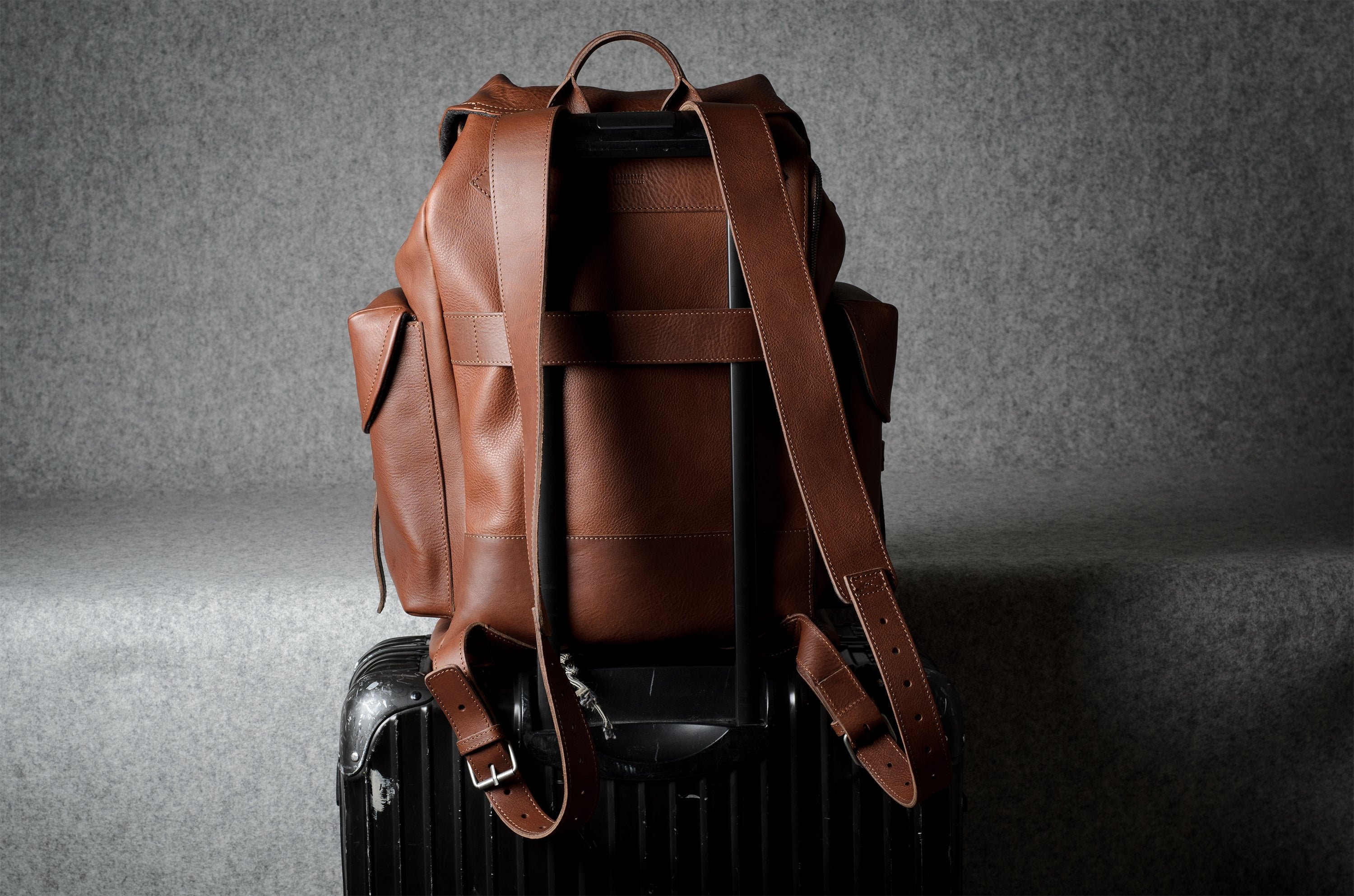 A brown leather backpack with wide adjustable shoulder straps, shown from the back resting on top of a black hard-shell suitcase against a textured gray studio background.
