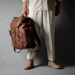 A person wearing cream-colored clothing and tan shoes holds a brown leather backpack against a textured gray studio backdrop.