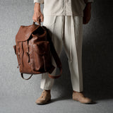 A person wearing cream-colored clothing and tan shoes holds a brown leather backpack against a textured gray studio backdrop.
