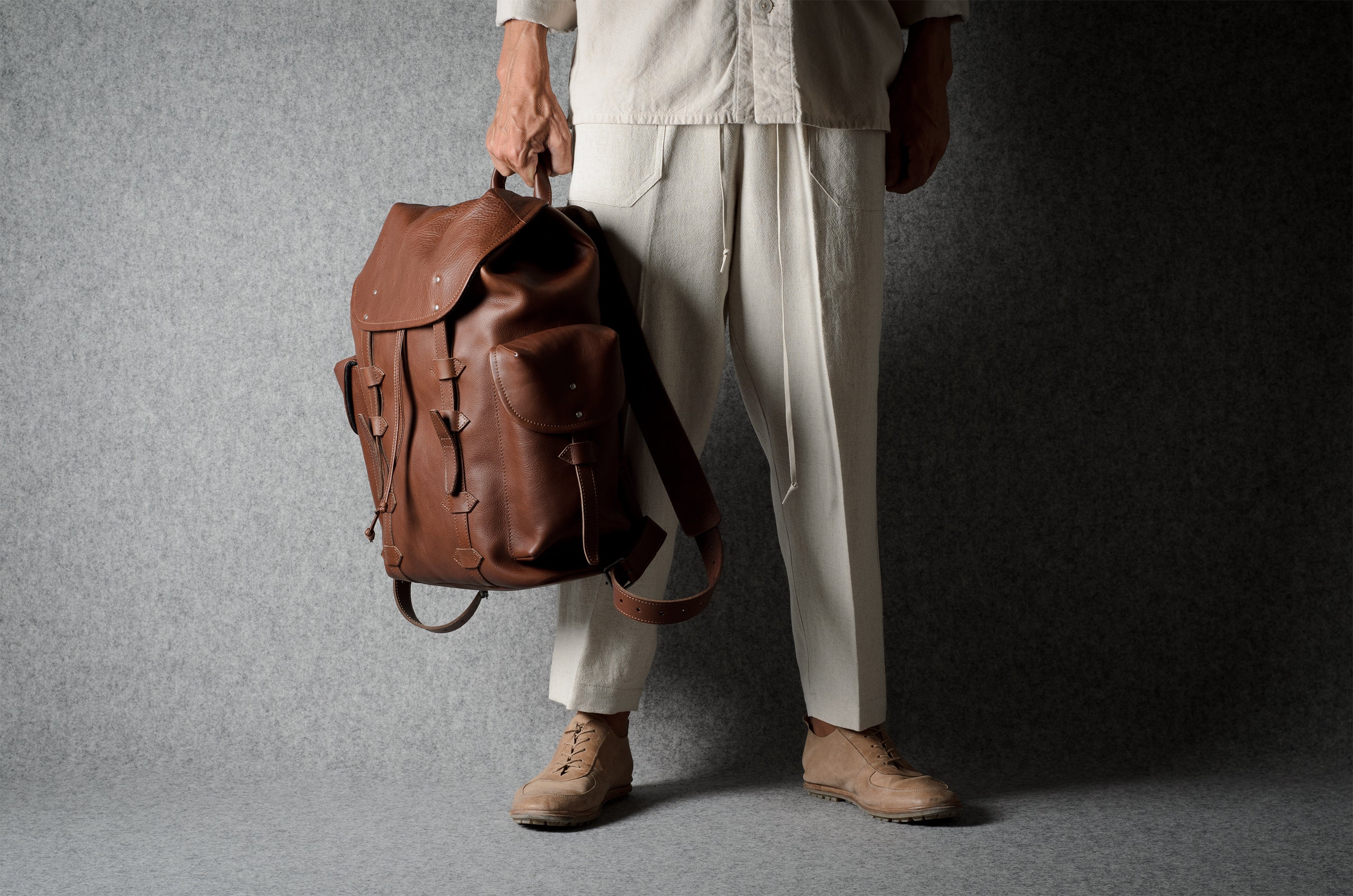 A person wearing cream-colored clothing and tan shoes holds a brown leather backpack against a textured gray studio backdrop.