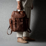 A person wearing off-white trousers and tan shoes holds a brown leather backpack against a dark gray studio backdrop.