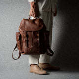 A person wearing off-white trousers and tan shoes holds a brown leather backpack against a dark gray studio backdrop.