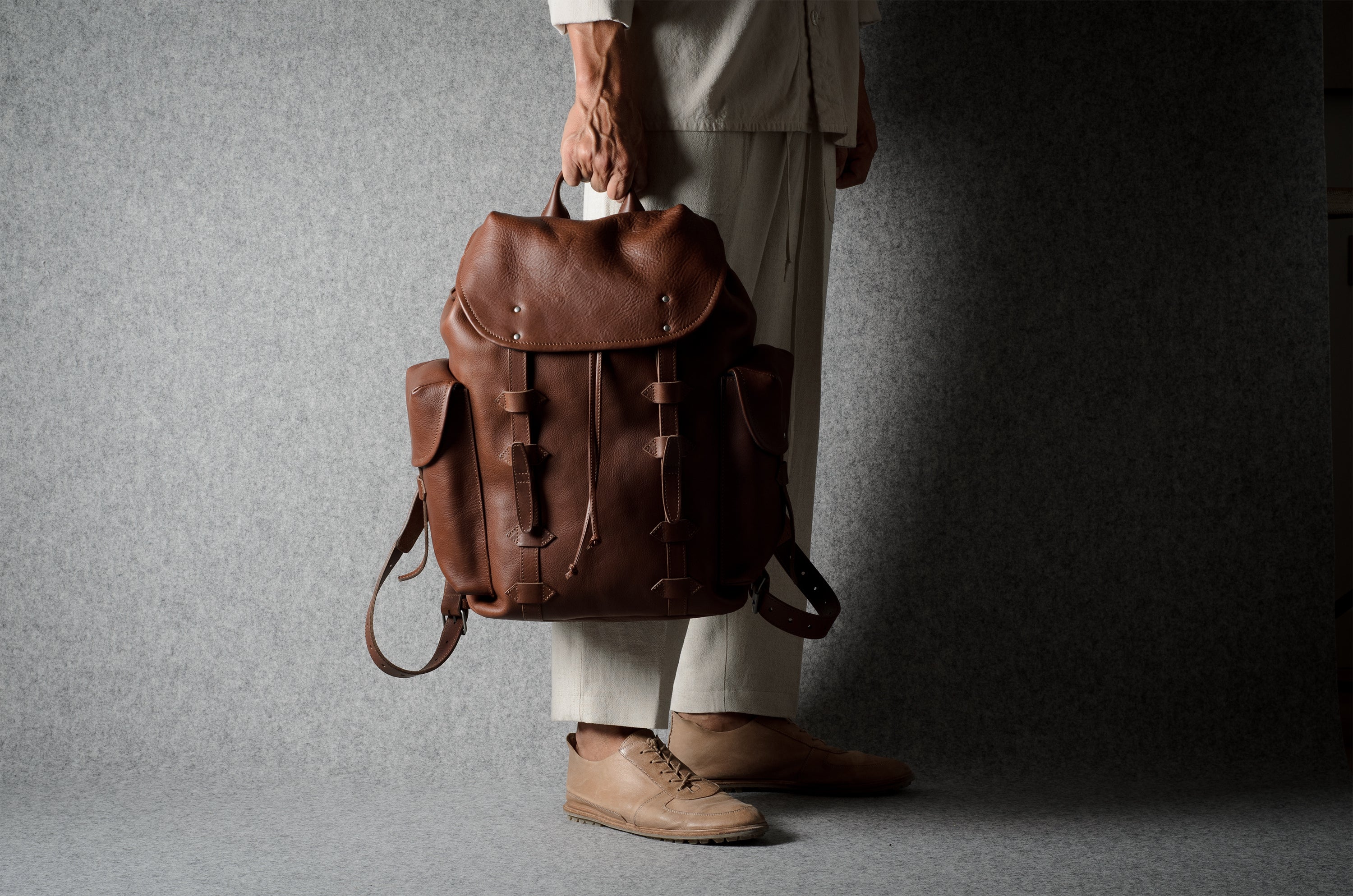 A person wearing off-white trousers and tan shoes holds a brown leather backpack against a dark gray studio backdrop.