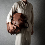 A person wearing a light beige linen outfit holds a brown leather duffel bag against a textured gray background.