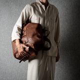 A person wearing a light beige linen outfit holds a brown leather duffel bag against a textured gray background.