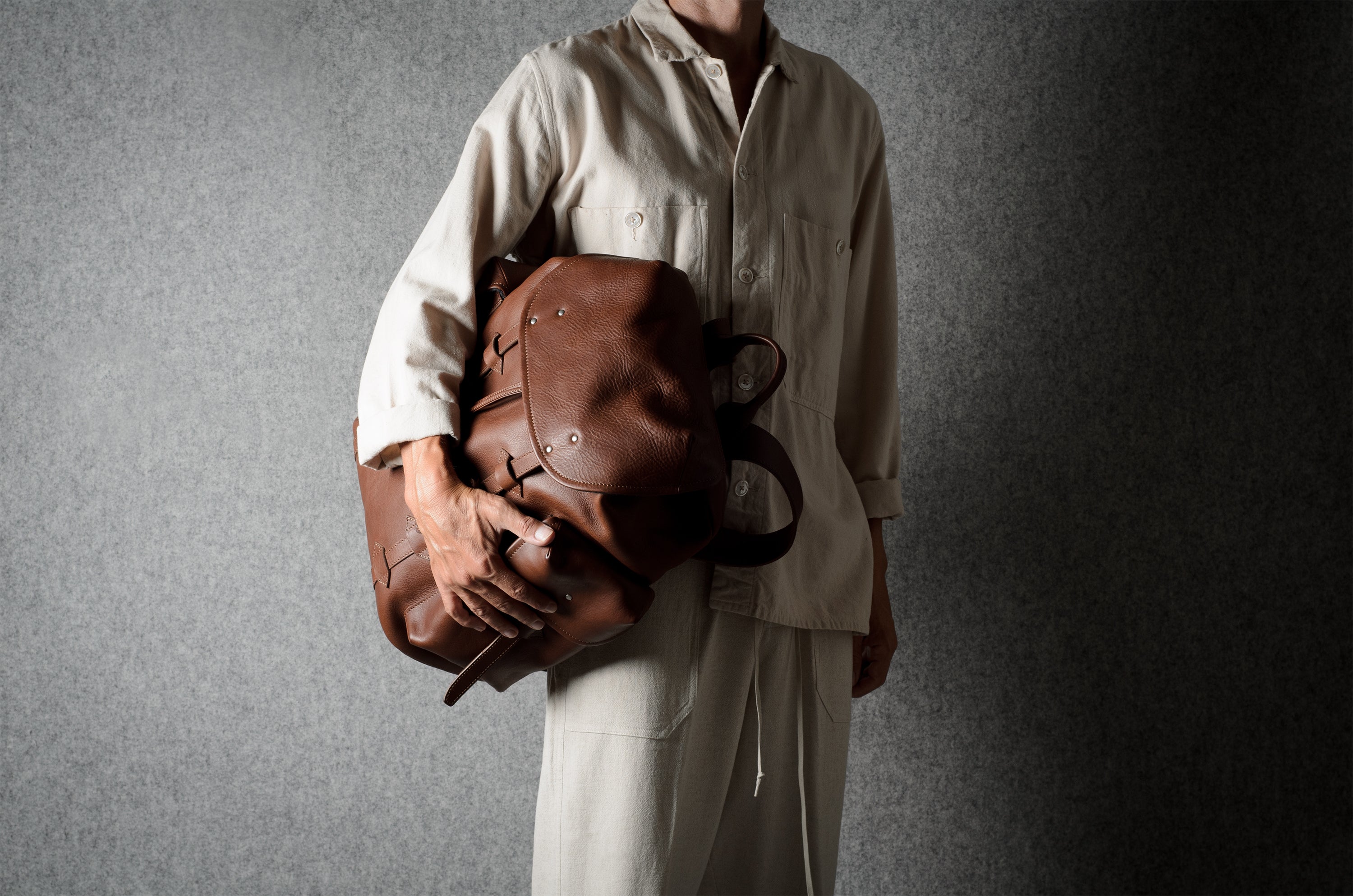 A person wearing a light beige linen outfit holds a brown leather duffel bag against a textured gray background.
