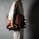 A person in a light beige shirt and trousers carries a brown leather backpack against a textured gray background.