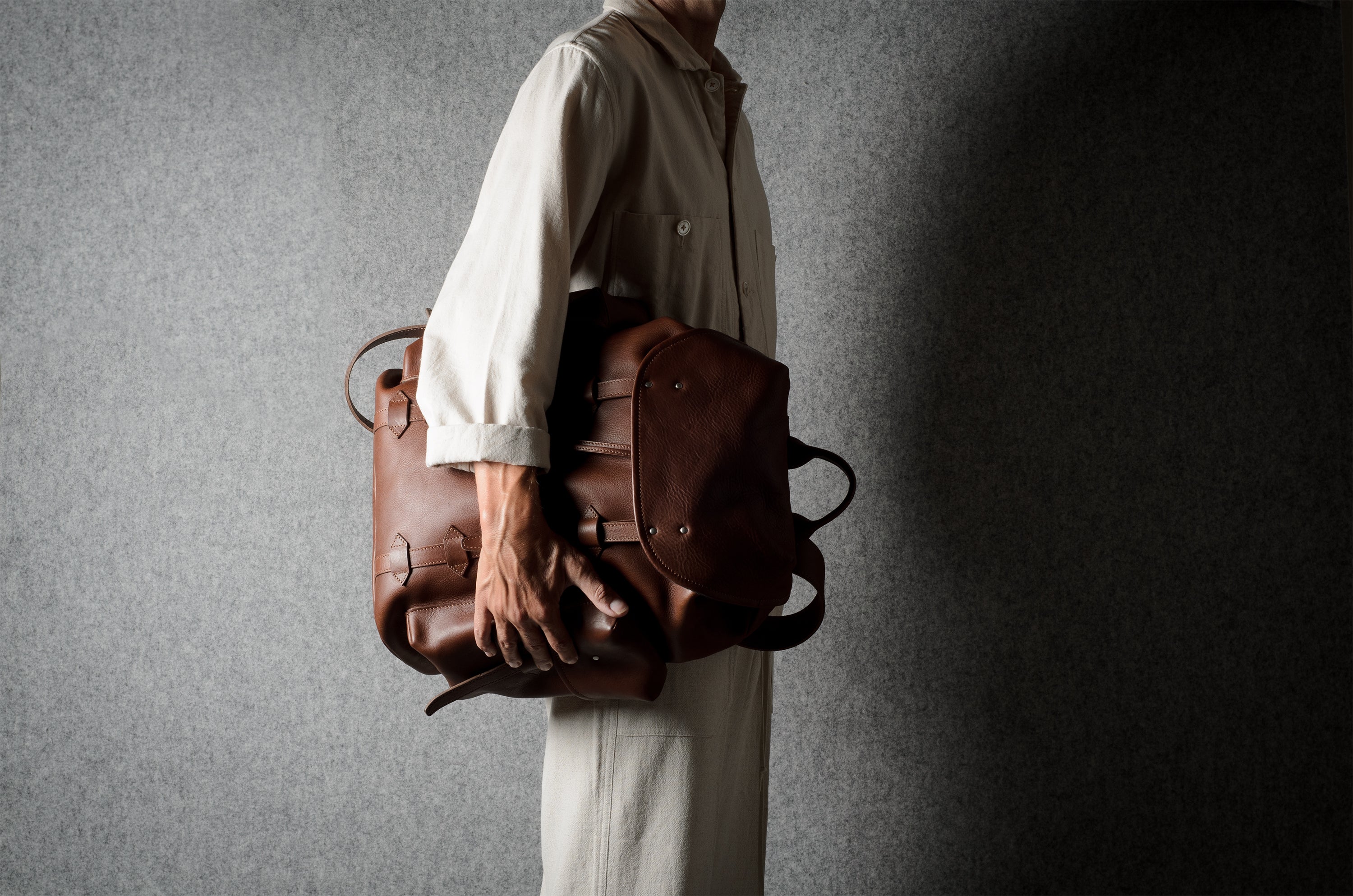 A person in a light beige shirt and trousers carries a brown leather backpack against a textured gray background.