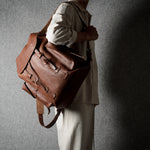 A person in a light beige outfit carries a large brown leather duffel bag against a textured gray wall with dramatic side lighting.