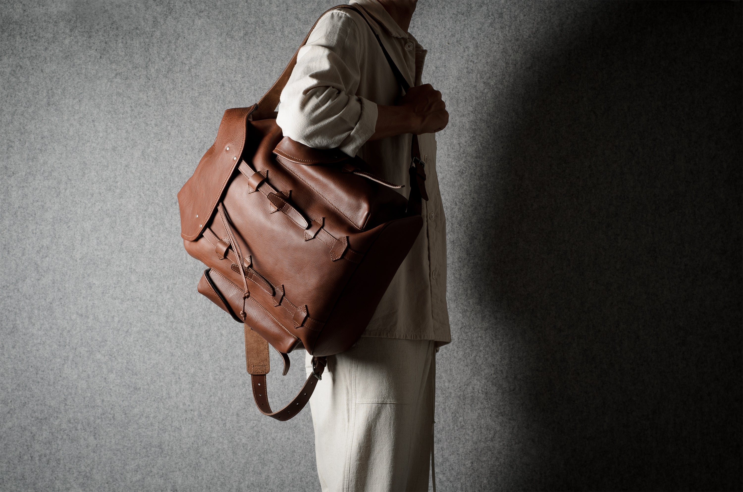 A person in a light beige outfit carries a large brown leather duffel bag against a textured gray wall with dramatic side lighting.